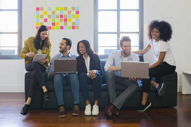 Happy colleagues sitting on couch using laptops - GIOF02700