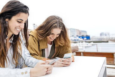 Zwei glückliche junge Frauen, die mit ihren Smartphones Nachrichten versenden - MGOF03420