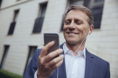 Porträt eines lächelnden reifen Geschäftsmannes im Freien mit Kopfhörern und Smartphone, lizenzfreies Stockfoto