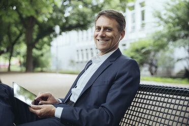 Lächelnder reifer Geschäftsmann sitzt mit Tablet und Kopfhörern auf einer Parkbank - MFF03616