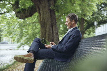 Lächelnder reifer Geschäftsmann sitzt mit Tablet und Kopfhörern auf einer Parkbank - MFF03617