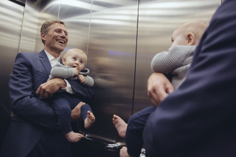Glücklicher reifer Geschäftsmann, der einen kleinen Jungen hält und in den Spiegel im Aufzug schaut, lizenzfreies Stockfoto