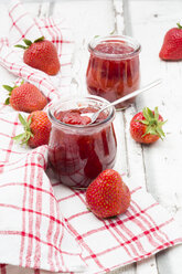 Zwei Gläser mit selbstgemachter Erdbeermarmelade, Küchenhandtuch und Erdbeeren auf weißem Holz - LVF06153