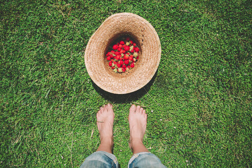Barfüßige Frau steht neben einem Strohhut voller Erdbeeren auf dem Rasen - GEMF01661
