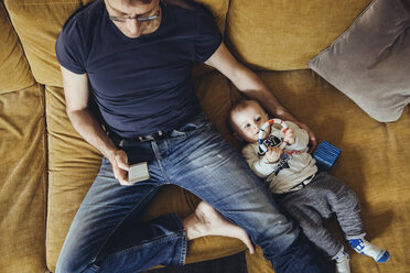 Baby boy lying on couch besides his father using smartphone - MFF03630