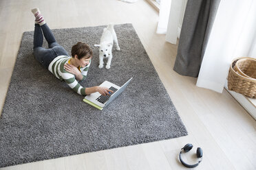 Woman lying on the floor in the living room using laptop while dog watching her - RBF05672