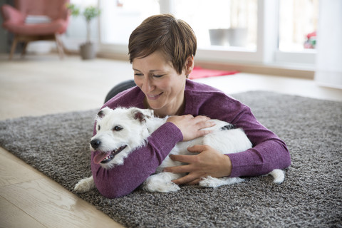 Frau liegt auf dem Teppich im Wohnzimmer und umarmt ihren Hund, lizenzfreies Stockfoto