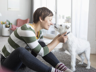 Lächelnde Frau, die auf dem Boden im Wohnzimmer sitzt und mit ihrem Hund kuschelt - RBF05696
