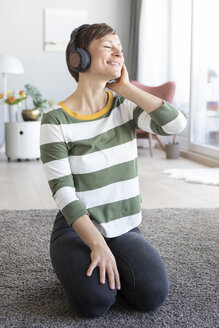 Porträt einer lächelnden Frau, die auf dem Boden im Wohnzimmer sitzt und mit Kopfhörern Musik hört - RBF05700