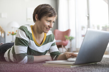 Smiling woman lying on the floor at home using laptop - RBF05708