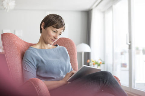 Porträt einer Frau, die zu Hause auf einem Sessel sitzt und ein Tablet benutzt, lizenzfreies Stockfoto