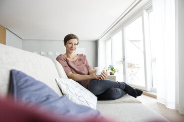 Porträt einer lächelnden Frau, die sich mit einer Tasse Kaffee auf der Couch entspannt - RBF05736