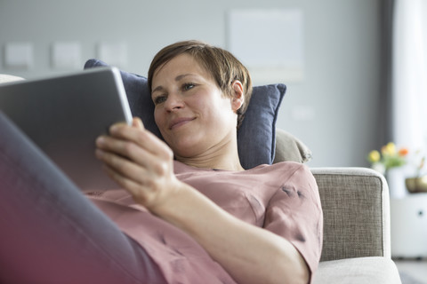 Porträt einer lächelnden Frau, die auf der Couch liegt und ein Tablet benutzt, lizenzfreies Stockfoto