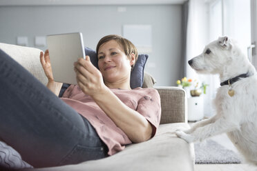 Porträt einer lächelnden Frau, die auf der Couch liegt und ein Tablet benutzt, während der Hund sie beobachtet - RBF05739