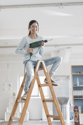 Junge Frau in ihrer neuen Wohnung, die auf einer Leiter sitzt und eine elektrische Bohrmaschine hält, lizenzfreies Stockfoto