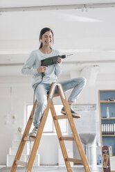 Young woman in her new flat sitting on ladder, holding electric drill - JOSF01086