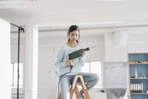 Junge Frau in ihrer neuen Wohnung, die auf einer Leiter sitzt und eine elektrische Bohrmaschine hält - JOSF01107