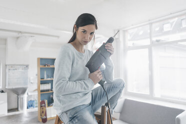 Junge Frau in ihrer neuen Wohnung, die auf einer Leiter sitzt und eine elektrische Bohrmaschine hält - JOSF01108