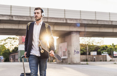 Young man on the move with skateboard, rolling suitcase and headphones - UUF10835