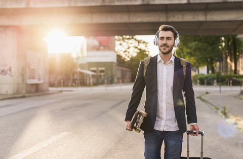 Young man on the move with skateboard, rolling suitcase and headphones stock photo