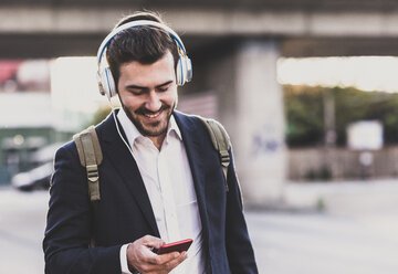 Lächelnder junger Mann, der Kopfhörer trägt und sein Mobiltelefon überprüft - UUF10838