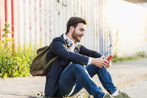 Lächelnder junger Mann, der auf dem Bürgersteig sitzt und sein Mobiltelefon überprüft - UUF10842