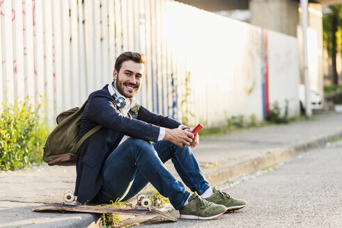 Lächelnder junger Mann sitzt auf dem Bürgersteig und hält ein Handy in der Hand - UUF10843