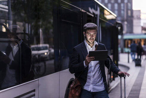 Junger Mann an der Bushaltestelle in der Stadt mit Kopfhörern und Tablet - UUF10858