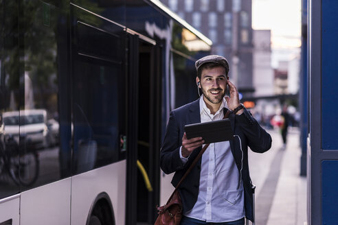 Junger Mann an der Bushaltestelle in der Stadt mit Kopfhörern und Tablet - UUF10859