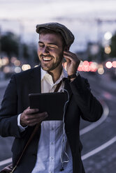 Happy young man in the city with earphones and tablet at dusk - UUF10861