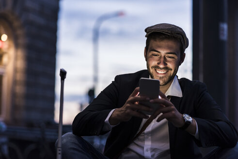 Lächelnder junger Mann in der Stadt, der am Abend sein Handy überprüft - UUF10862
