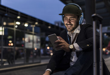 Junger Mann in der Stadt mit Kopfhörern und Mobiltelefon am Abend - UUF10863
