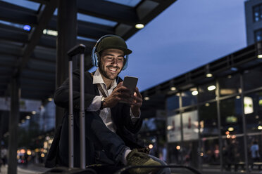 Junger Mann in der Stadt mit Kopfhörern und Mobiltelefon am Abend - UUF10864