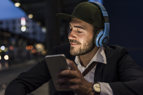 Junger Mann in der Stadt mit Kopfhörern und Mobiltelefon am Abend - UUF10866