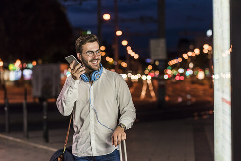 Junger Mann in der Stadt mit Kopfhörern und Mobiltelefon bei Nacht - UUF10870