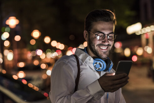 Lächelnder junger Mann in der Stadt, der nachts sein Mobiltelefon überprüft - UUF10872