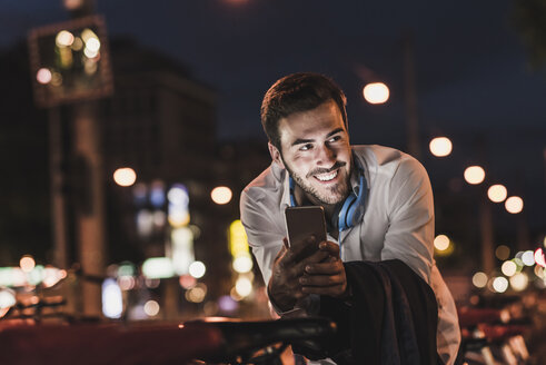 Lächelnder junger Mann in der Stadt mit Mobiltelefon bei Nacht - UUF10874