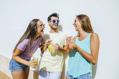 Laughing friends holding refreshing drinks in front of white wall - KIJF01521