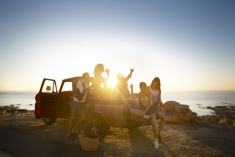 Glückliche junge Leute vor einem Pick-up-Truck an der Küste bei Sonnenuntergang, lizenzfreies Stockfoto