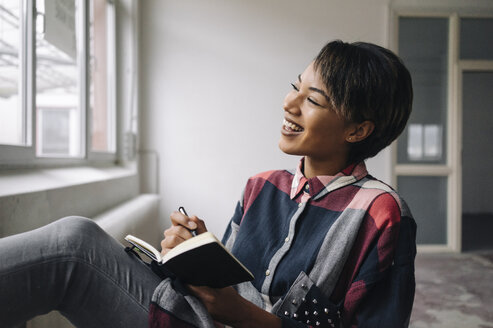 Smiling woman sitting at the window with notebook - KNSF01520