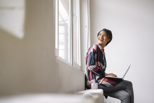 Smiling woman sitting at the window using laptop - KNSF01522