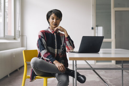 Confident young woman sitting on chair with laptop on table - KNSF01533