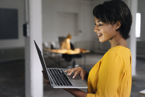 Smiling woman using laptop in empty office - KNSF01537