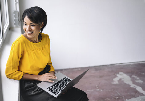 Smiling woman sitting at the window using laptop - KNSF01539
