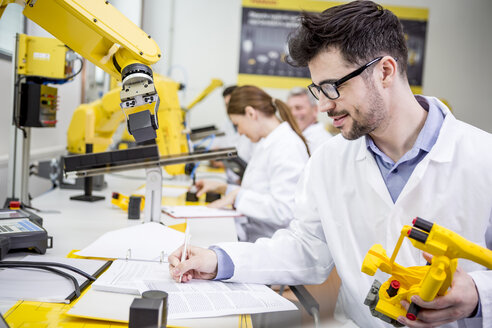 Engineer in factory holding model of an industrial robot taking notes on clipboard - WESTF23419