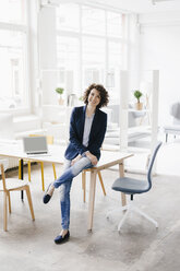 Businesswoman in office sitting on desk, looking confident - KNSF01551