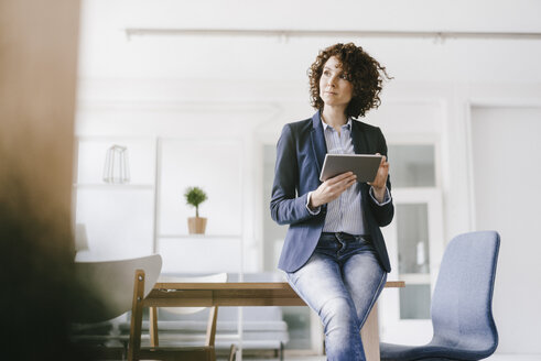 Businesswoman in office sitting on desk, using digital tablet - KNSF01553