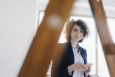 Geschäftsfrau steht an einer Leiter im Büro und hält ein Smartphone - KNSF01558
