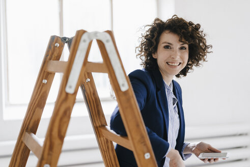 Businesswoman standing by ladder in office, holding smartphone - KNSF01560