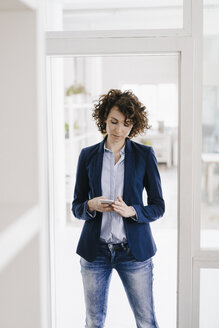 Geschäftsfrau, die in der Bürotür steht und ein Smartphone benutzt - KNSF01577